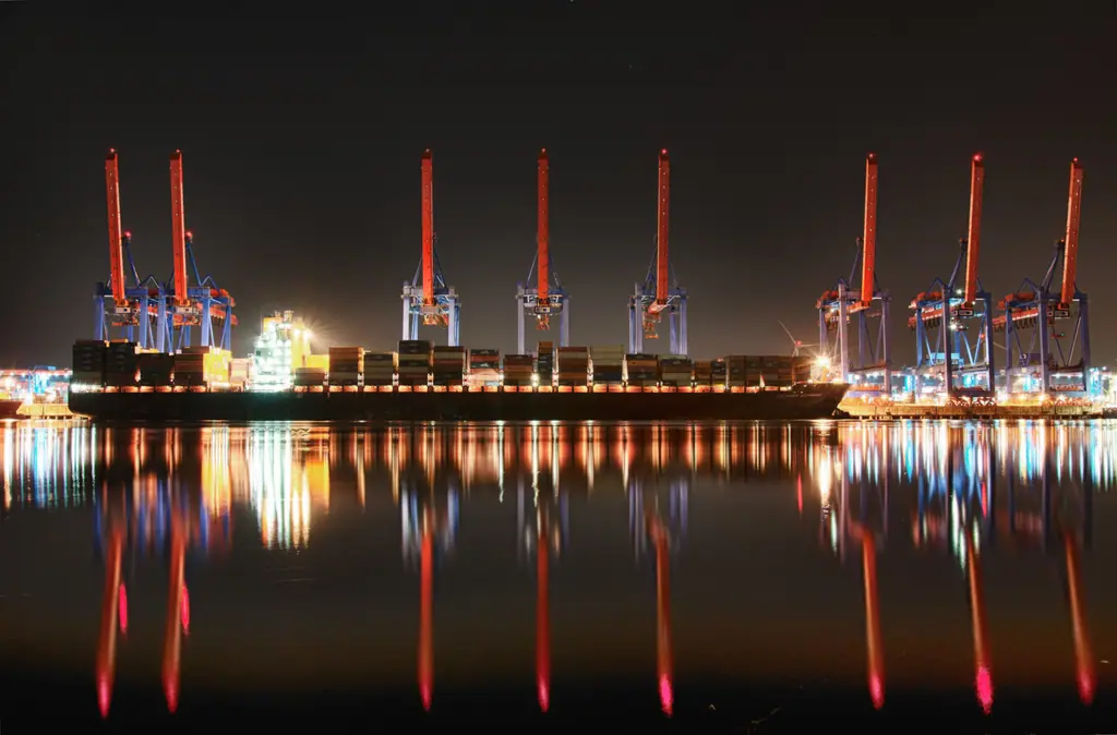 A container ship berthed at a port terminal at night, its lights reflected in the water